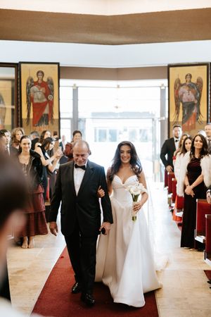 Bride in white off-shoulder gown walks down red carpet aisle with father in Orthodox church.