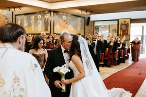 Bride in white veil carrying calla lily bouquet walks down church aisle with father during wedding ceremony.
