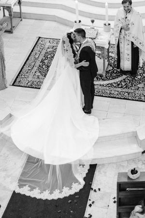 Black and white photo of bride and groom sharing first kiss at Orthodox church altar with priest.