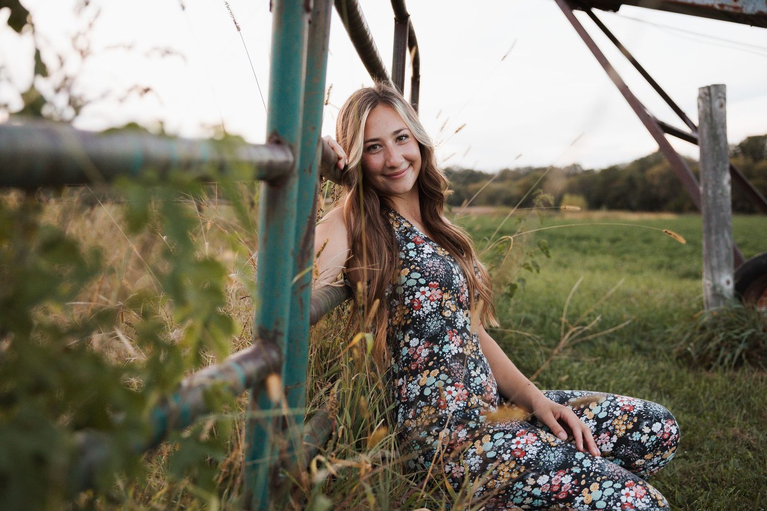 A person in a floral dress poses in a golden field at sunset with a rustic fence in the background.