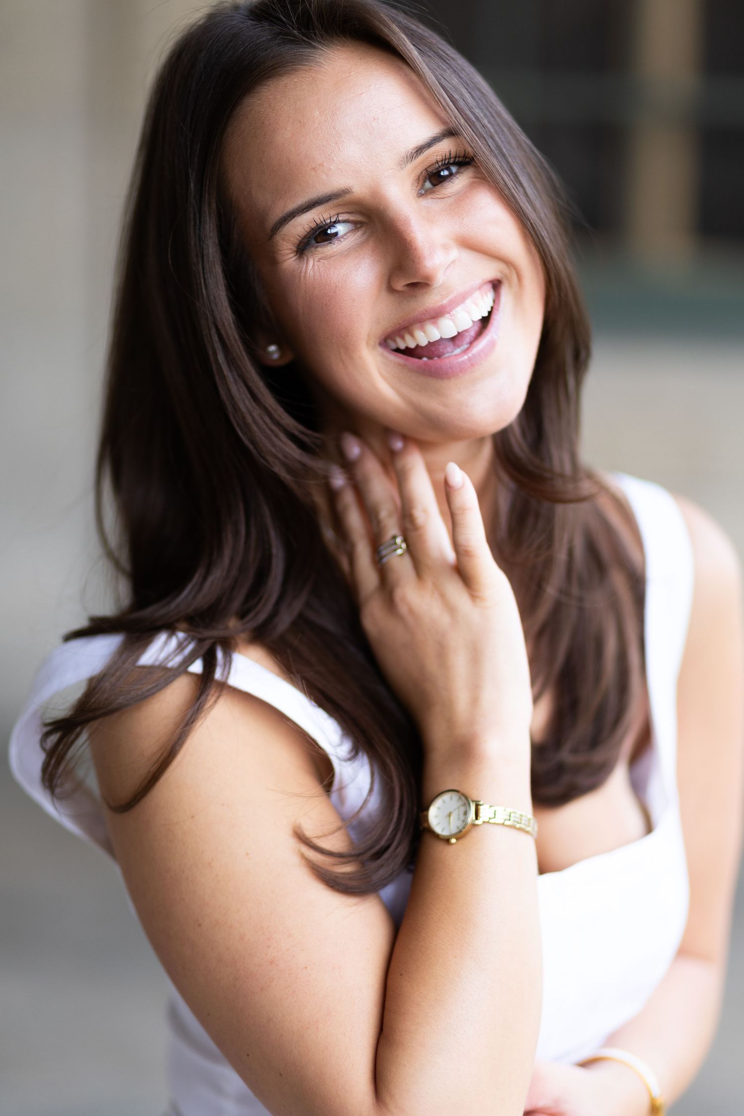 Smiling brunette woman in white top wearing a gold watch, posing with hand near chin.