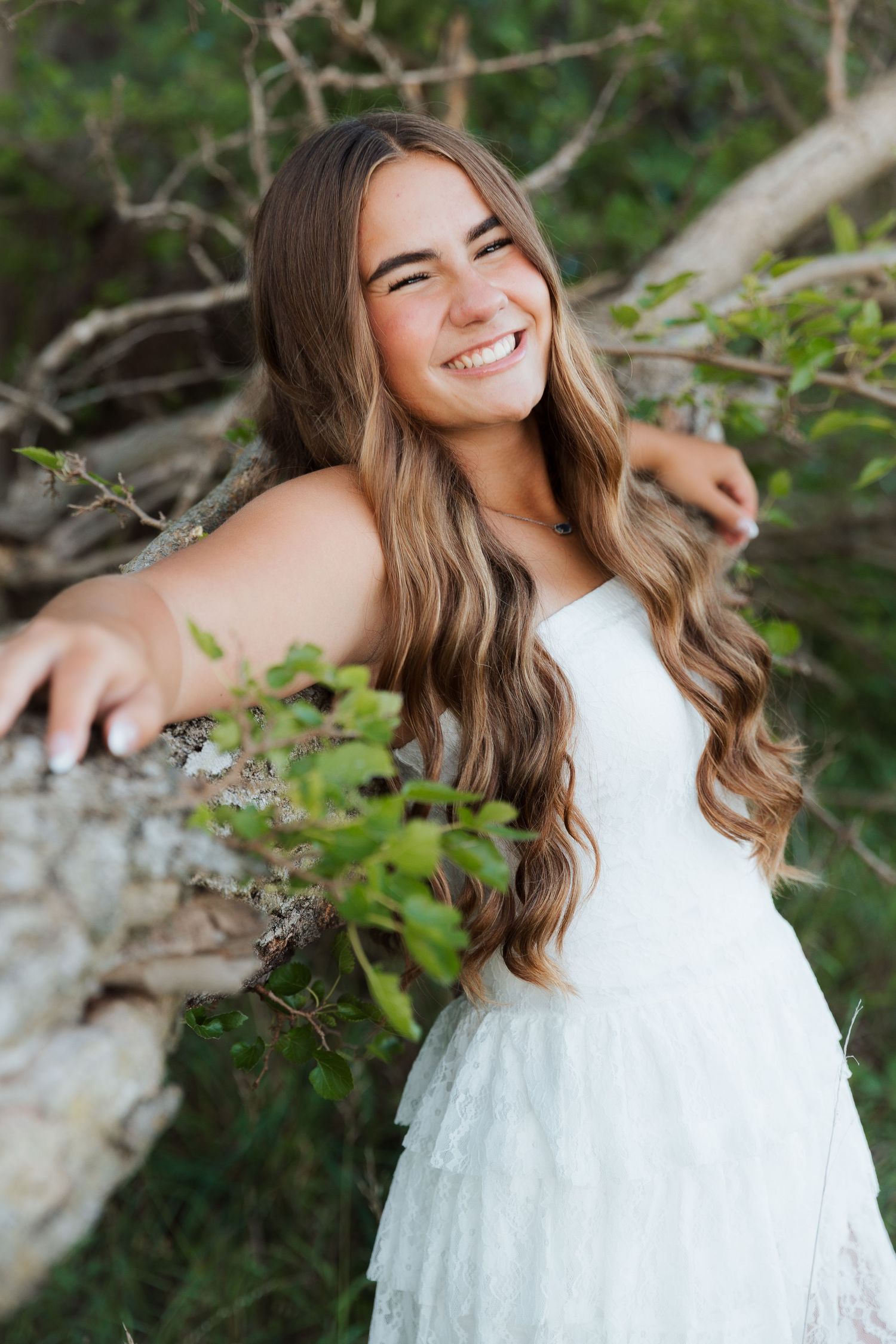 Young woman in white dress smiling outdoors, leaning against a tree with long wavy hair and green foliage background.