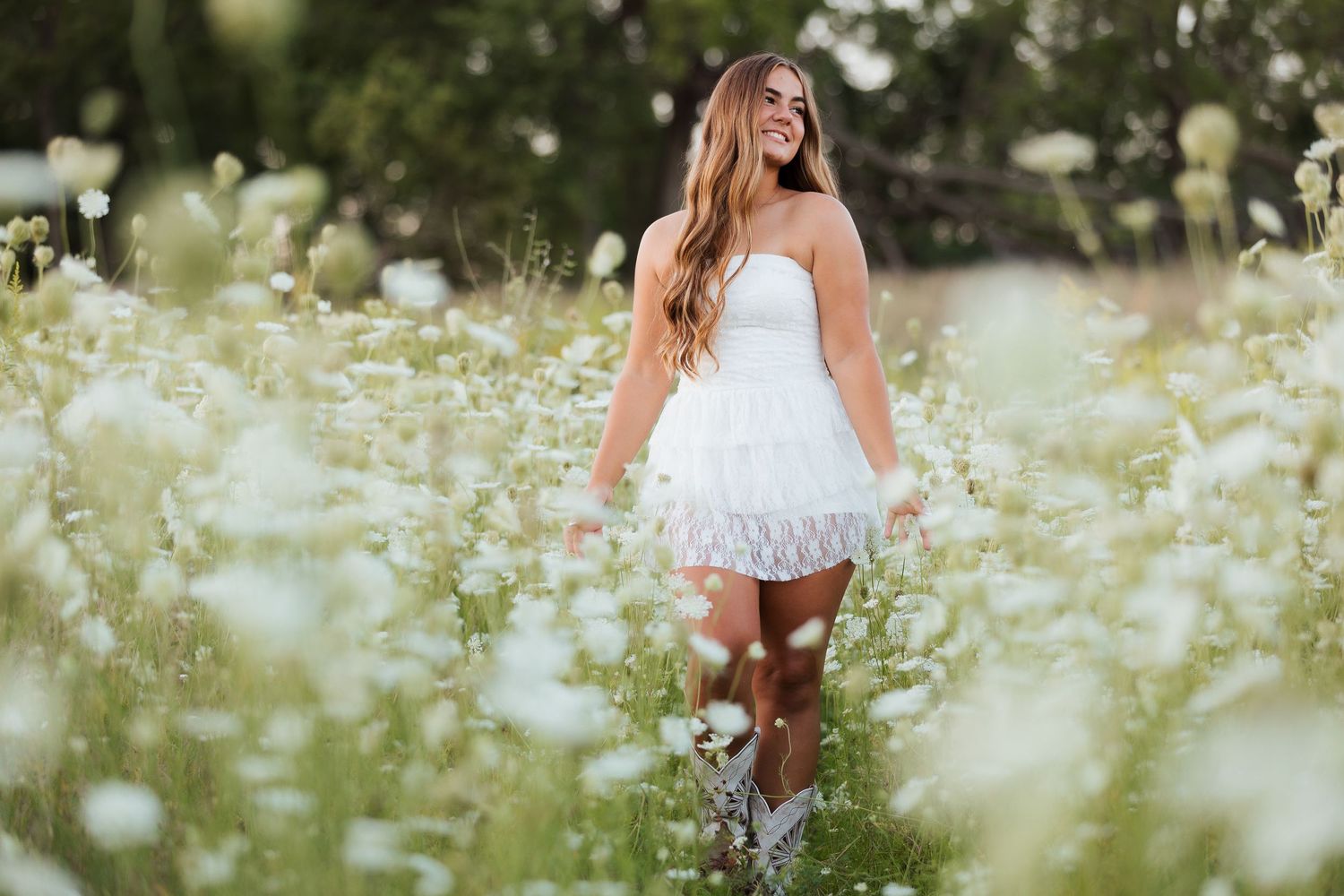 Young woman in white dress walking through a field of white wildflowers, smiling joyfully on a sunny day.