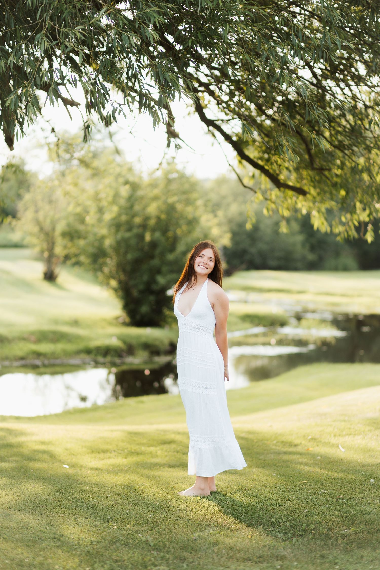 Young woman in white maxi dress smiling in lush green park with pond and willow tree backdrop.