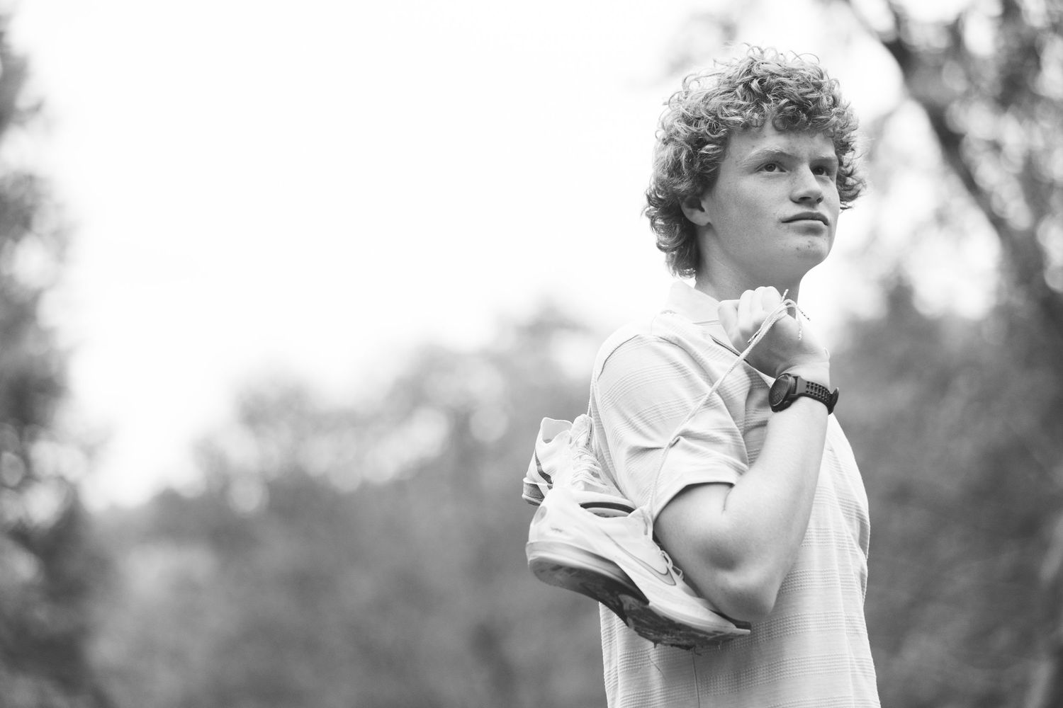 Black and white photo of a young man with curly hair holding sneakers over his shoulder outdoors.