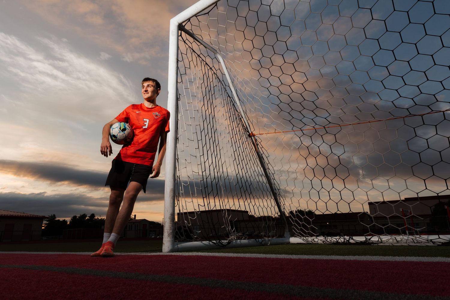 Soccer player in red jersey holding ball beside goal post at sunset on a track field.