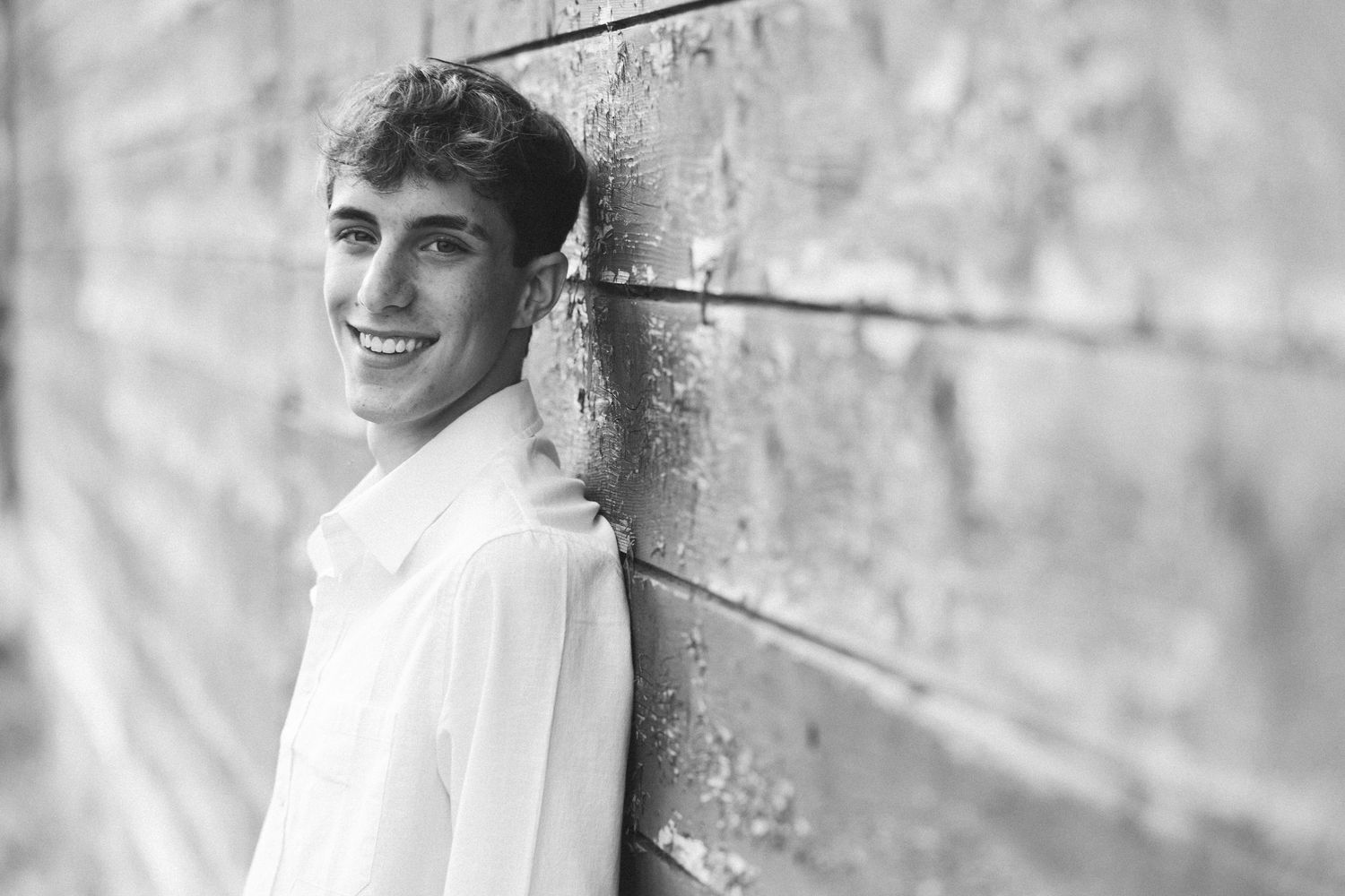 Black and white portrait of a smiling young man in a white shirt leaning against a stone wall.