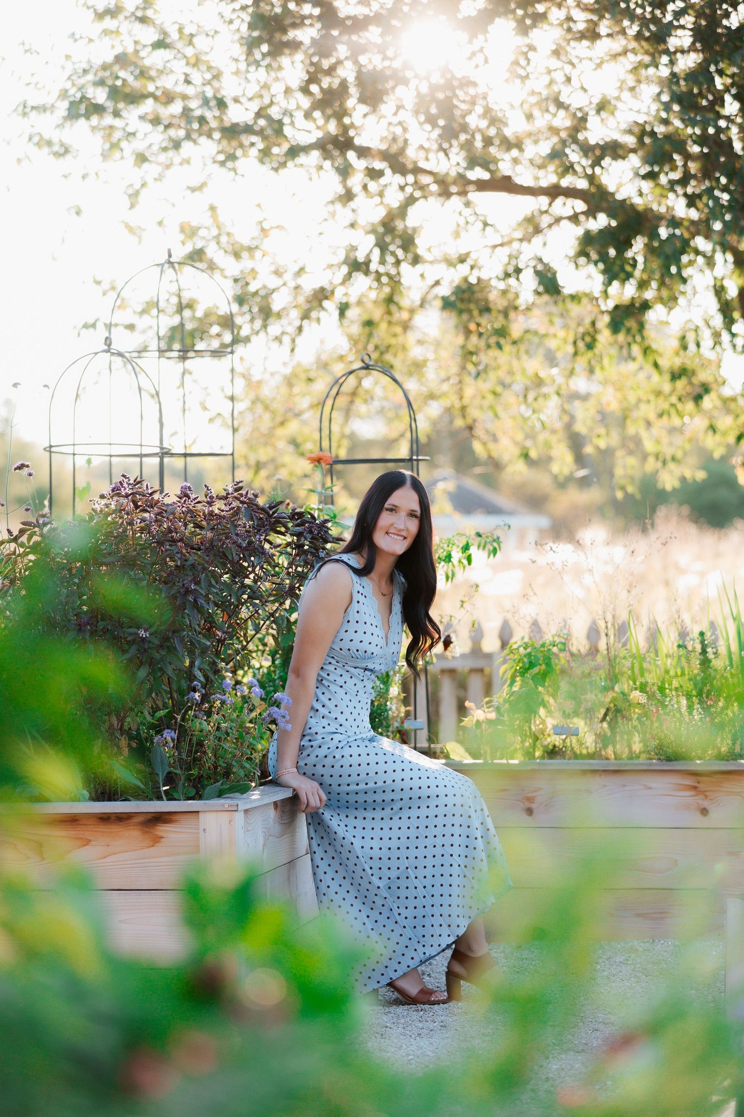 Woman in polka dot maxi dress sitting on raised garden bed surrounded by lush greenery in golden sunlight.