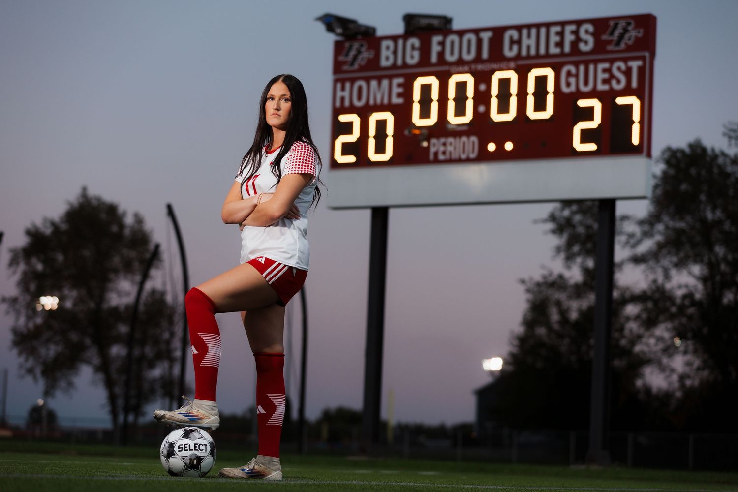 Female soccer player poses confidently in red and white uniform beside a ball in front of Big Foot Chiefs scoreboard at dusk.