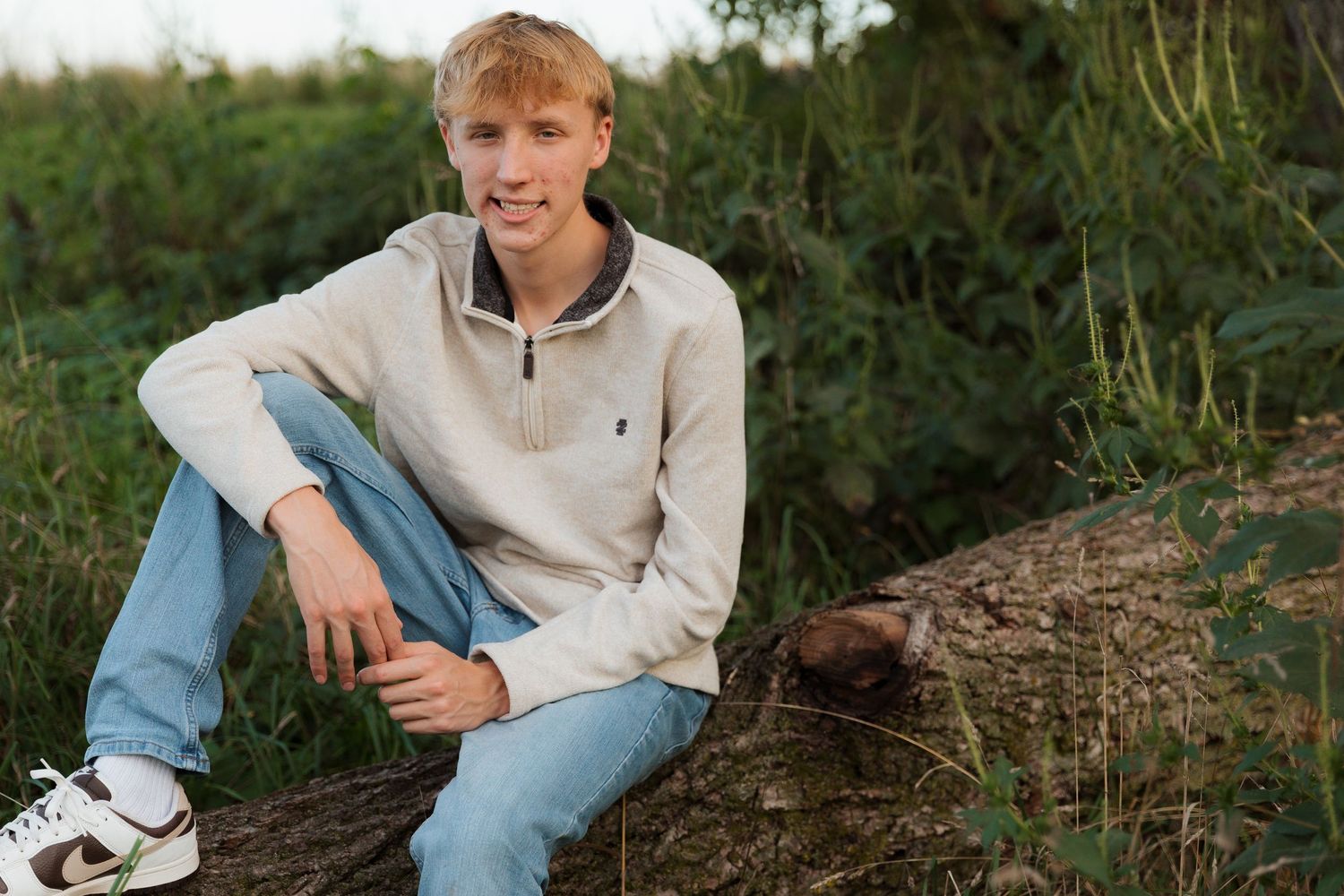 Young man in beige sweater and jeans sitting on a log outdoors surrounded by lush green vegetation.