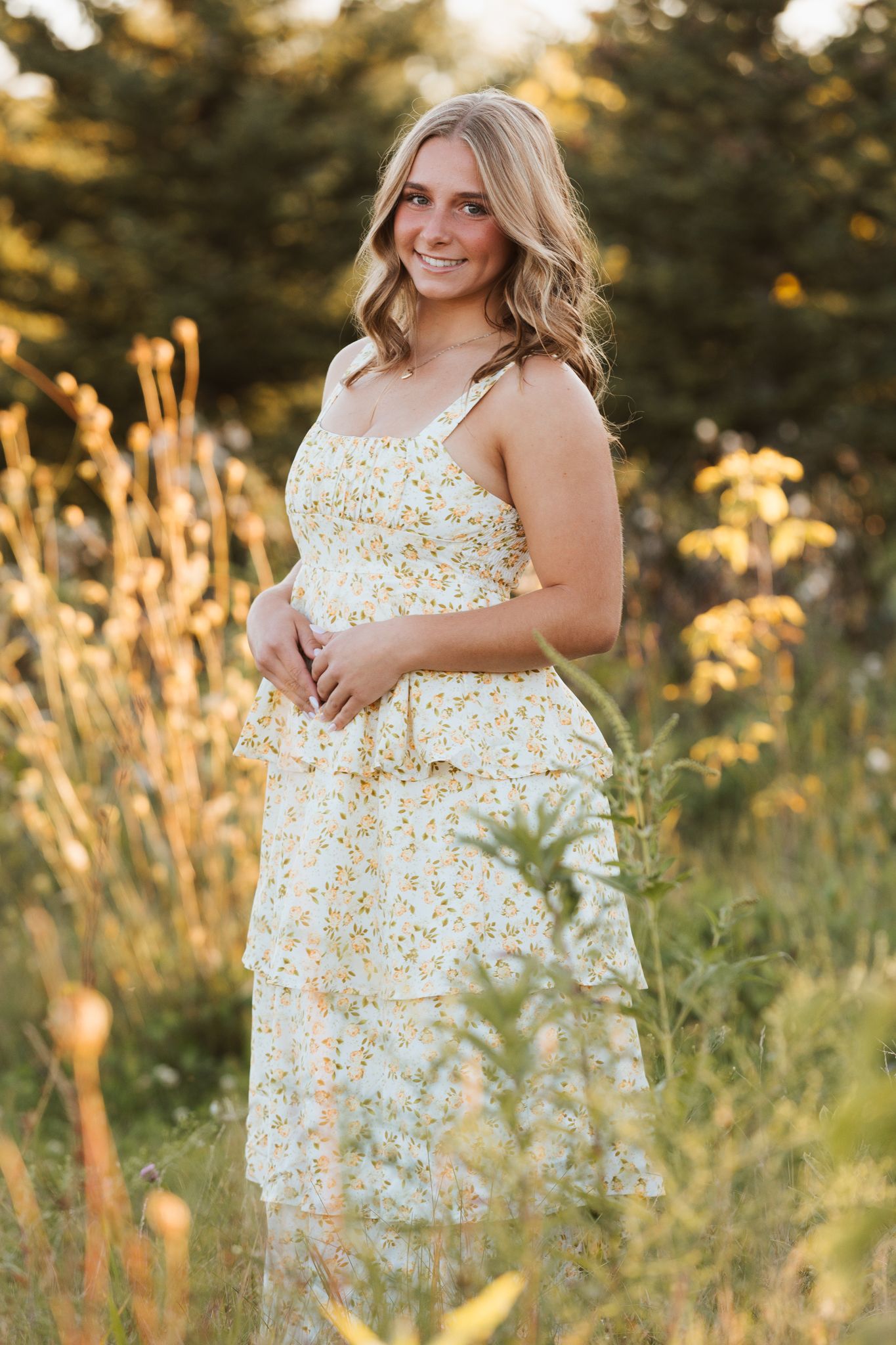 Woman in floral sundress poses outdoors in golden sunlit field with tall grass at sunset.
