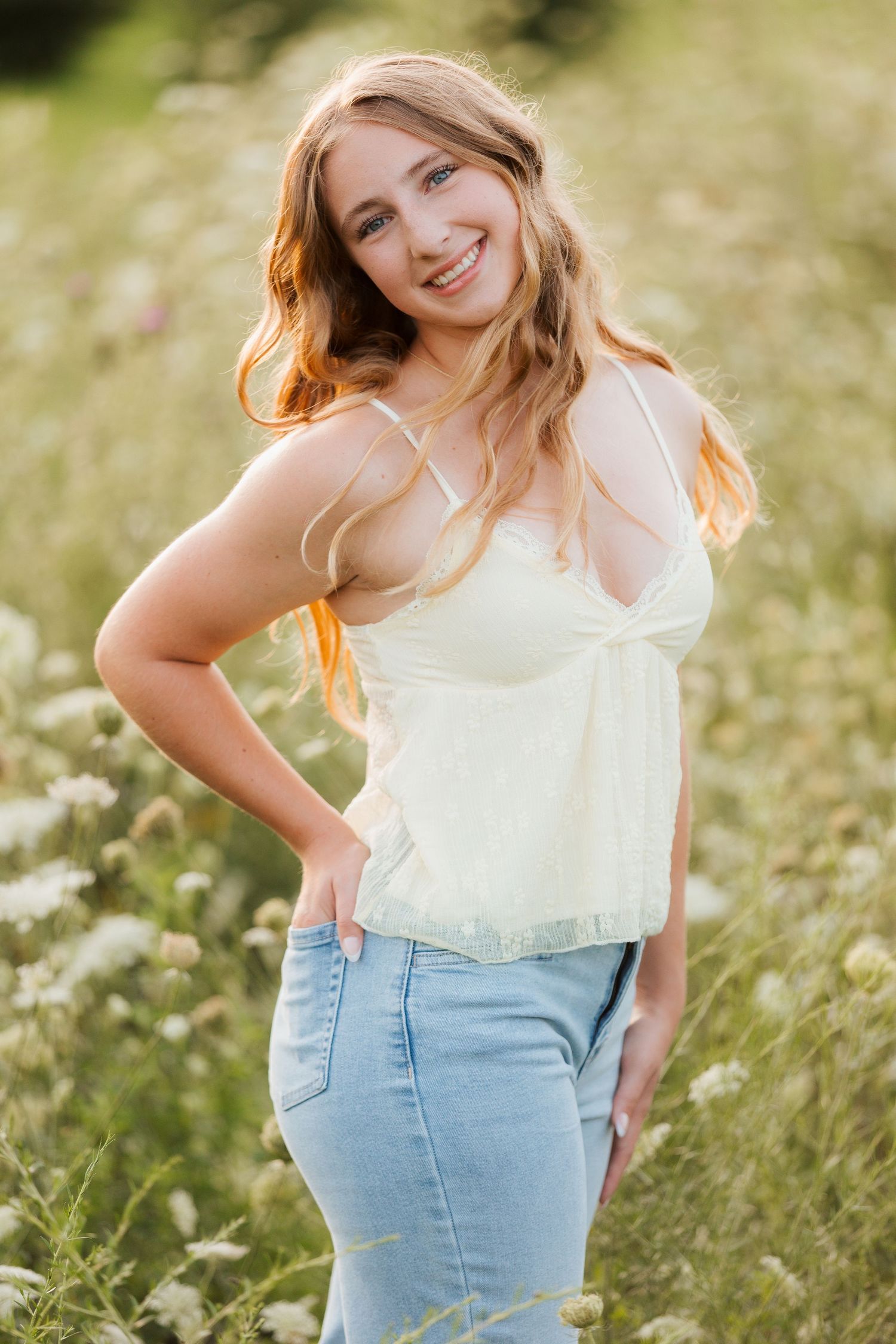 Smiling young woman with blonde hair in white camisole and jeans posing in a sunny wildflower field.