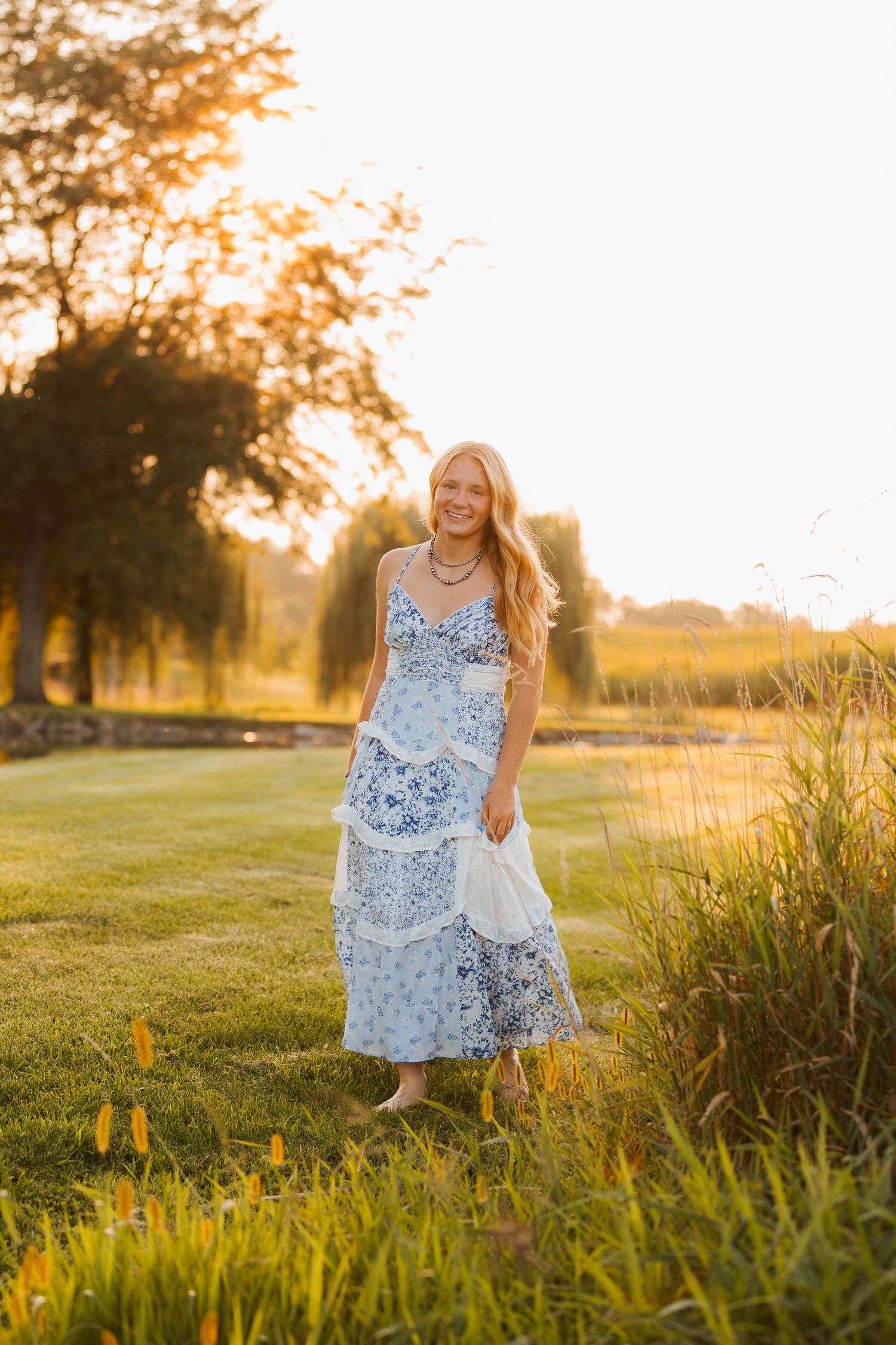 Young woman in blue floral maxi dress standing in golden hour sunlight on lush green grass outdoors.