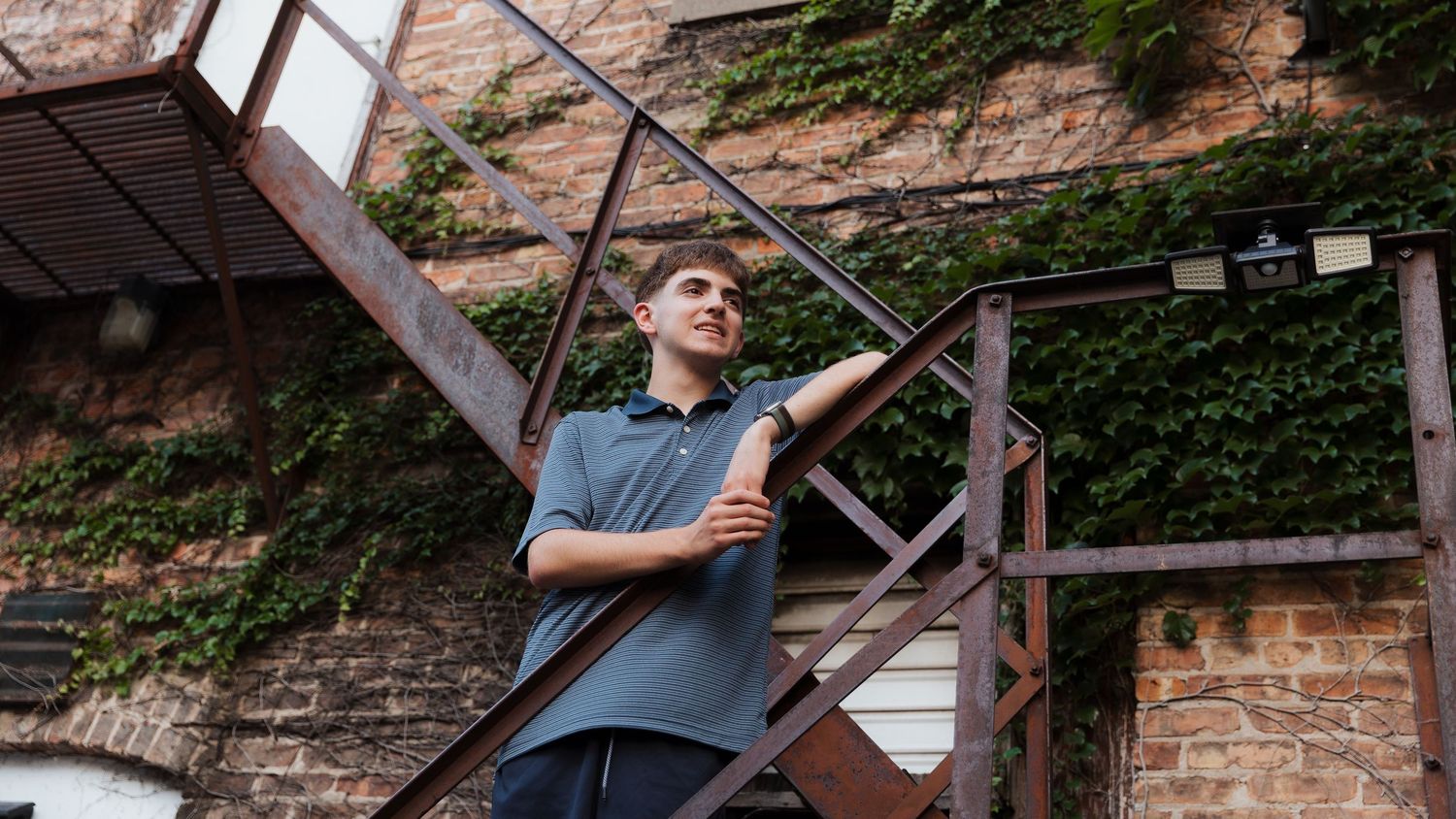 Young man in gray polo shirt leaning on rustic brick building staircase surrounded by lush green ivy.