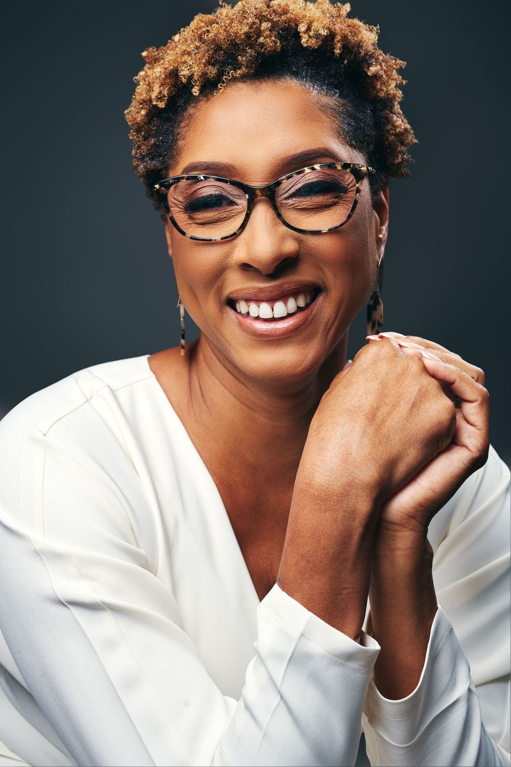 Woman with short curly hair and glasses smiles warmly in white blouse against dark backdrop.