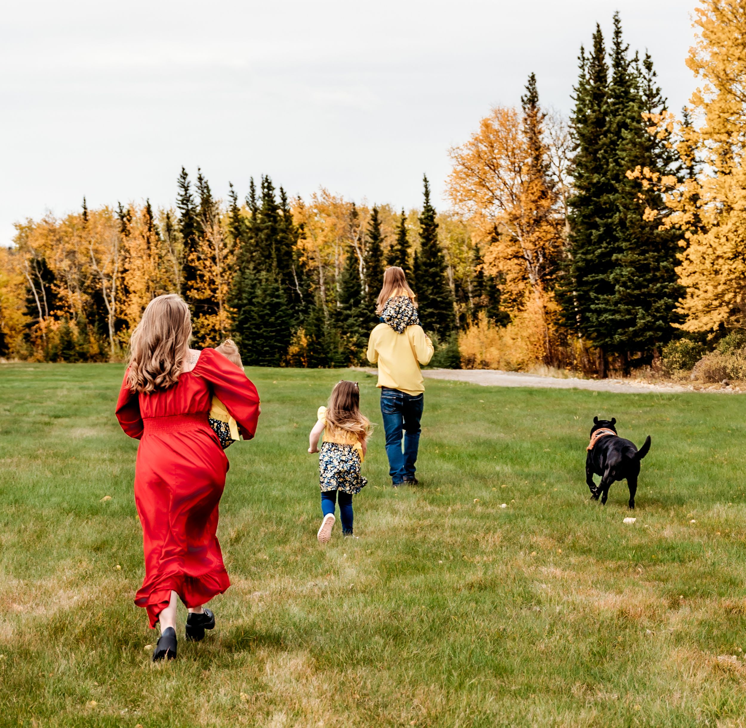 Family and dog running through grass field with autumn trees in background.