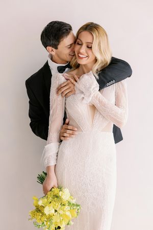 A couple embraces in wedding attire with the bride holding a yellow bouquet against a white background.