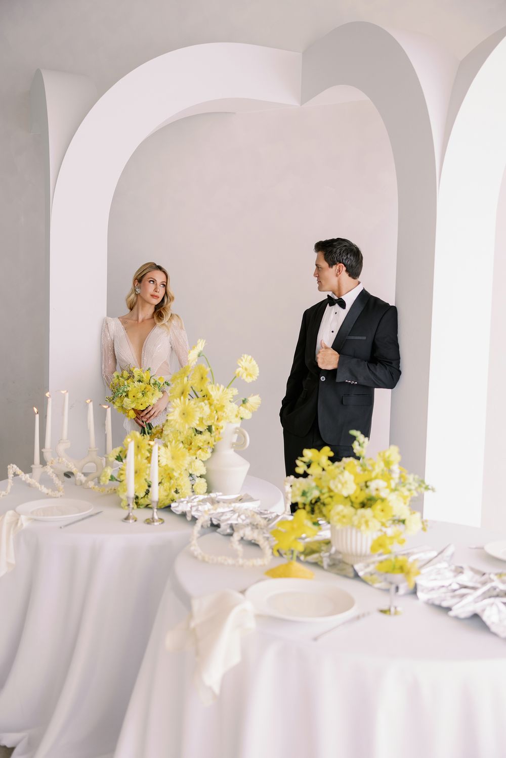 Bride and groom at elegant white wedding reception with yellow floral arrangements and candles under white archway.
