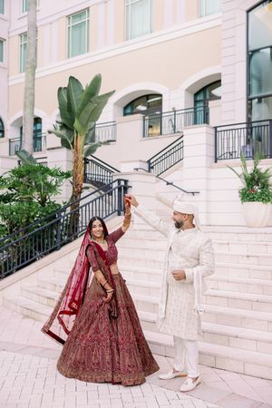 Couple in traditional South Asian wedding attire dancing on elegant white marble steps with palm trees and architecture.