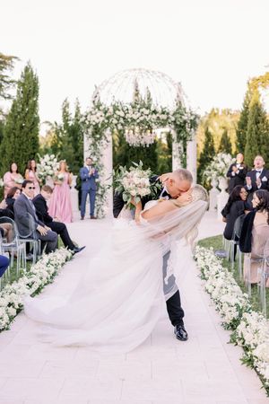 Romantic wedding couple embraces and dips under floral arch at outdoor ceremony with guests seated on white chairs.