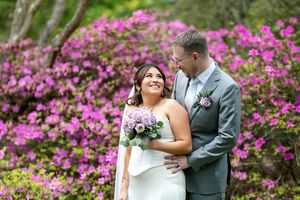 Wedding couple embracing before vibrant pink azalea bushes, bride holding purple bouquet, groom in gray suit.