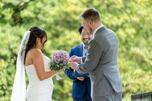 Groom placing wedding ring on brides finger during outdoor ceremony with smiling officiant.