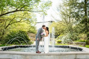 Wedding couple sharing a kiss by a garden fountain, bride in white gown holding bouquet, groom in gray suit.
