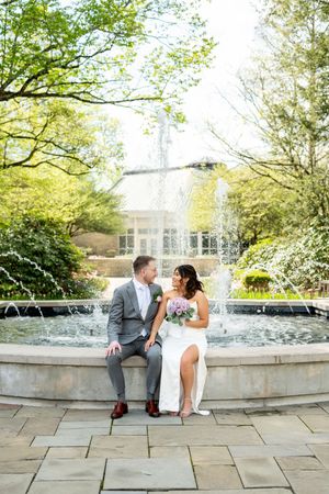 Bride and groom sitting together on fountain edge in lush garden, sharing a romantic glance on wedding day.