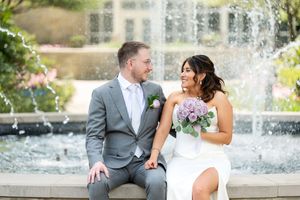 Bride in strapless white gown and groom in gray suit sitting by fountain, holding hands with purple bouquet.