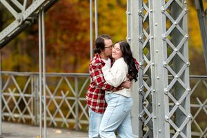 Romantic couple embracing and smiling on a metal bridge surrounded by fall foliage.
