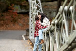 Couple sharing a kiss on an outdoor bridge with colorful autumn trees in background.