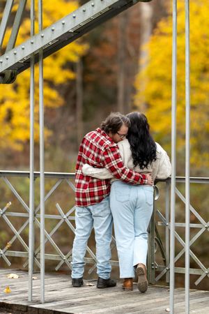 Couple hugging closely on a wooden bridge with vibrant yellow fall foliage behind them.