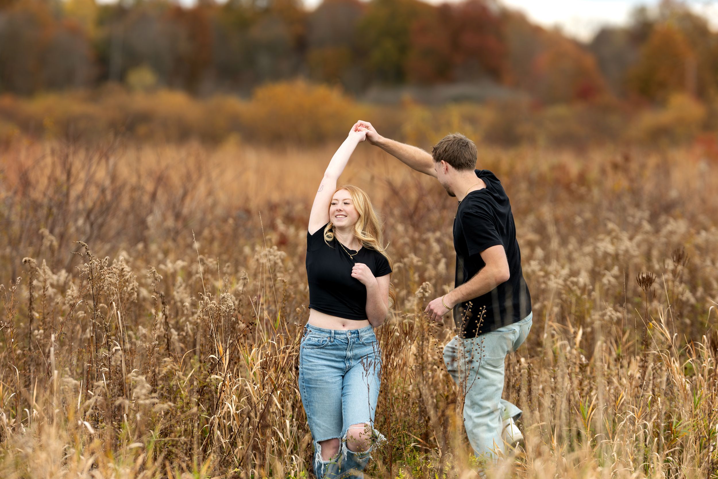 Man spinning woman in black crop top through tall golden grass in autumn field.