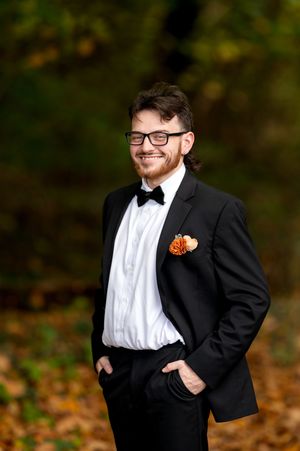 Smiling groom in black tuxedo with orange boutonniere posing outdoors in autumn setting.