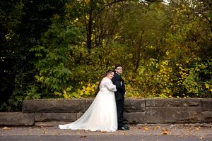 Bride and groom embracing near stone wall with lush green and yellow autumn trees.