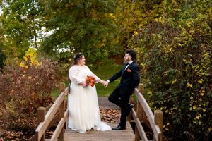 Bride and groom holding hands on wooden bridge surrounded by vibrant fall foliage.