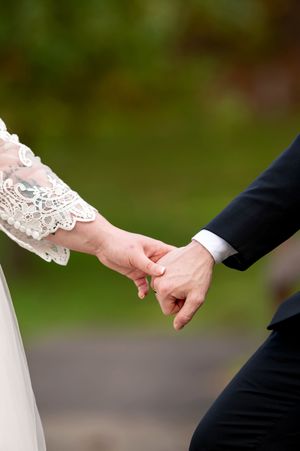 Close-up of bride and groom holding hands, bride wearing lace sleeve wedding dress outdoors.