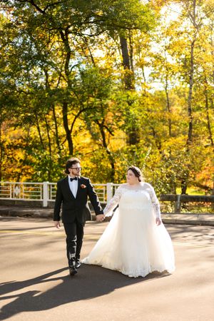 Newlyweds walk hand in hand down a sunlit path with golden fall trees in the background.