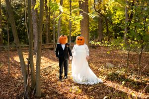 Playful wedding couple wearing carved jack-o-lantern pumpkin heads in an autumn forest setting.
