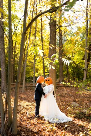 Halloween-themed bride and groom with pumpkin heads embrace among colorful fall foliage in the woods.