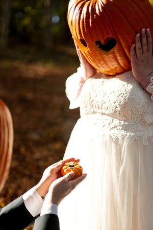 Groom presents bride wearing a pumpkin head with a tiny decorative pumpkin in a sunlit forest.