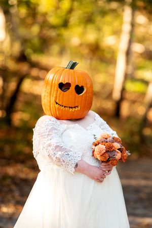 Bride in white lace gown holds orange bouquet while wearing a heart-eyed carved pumpkin on her head.