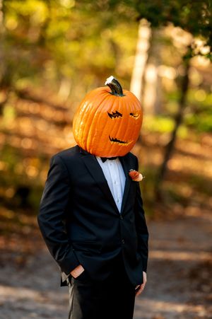 Groom in black tuxedo wearing a carved jack-o-lantern pumpkin head stands in an autumn forest.