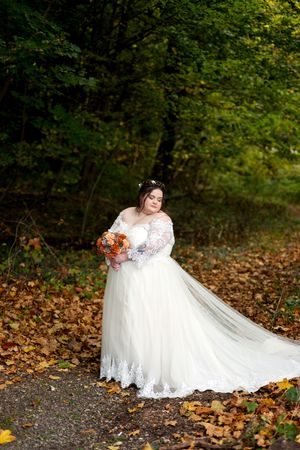 Bride in white lace gown holding orange bouquet standing among colorful autumn forest leaves.