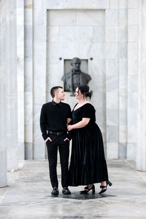 Couple dressed in elegant black attire standing together in a grand marble hall with a bust sculpture.