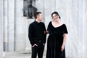 Couple in matching black outfits smiling together in a classic marble columned architectural setting.