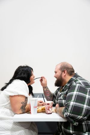 Tattooed couple sharing a fun moment feeding each other at a fast food restaurant engagement shoot.