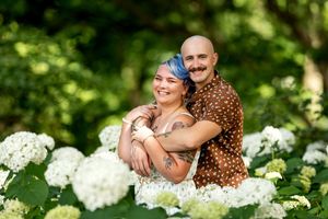 Smiling engaged couple embracing among white hydrangea blooms in a lush green garden setting.