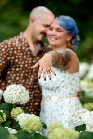 Engaged couple showing off ring among white hydrangeas, woman extending tattooed hand toward camera.