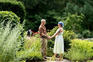 Engaged couple holding hands and facing each other on a garden path surrounded by colorful flowers.